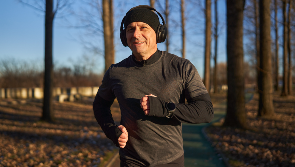 Man jogging outdoors in a park, wearing headphones and winter running gear, smiling while exercising on a sunny day.