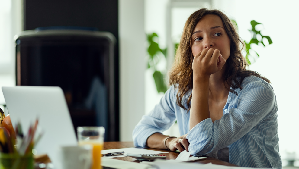 Woman at desk, looking thoughtful and concerned, working from home with a laptop and papers.