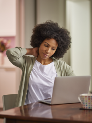 Woman sitting at a table, looking at her laptop and massaging her neck to relieve discomfort, wearing a casual green sweater and white shirt.