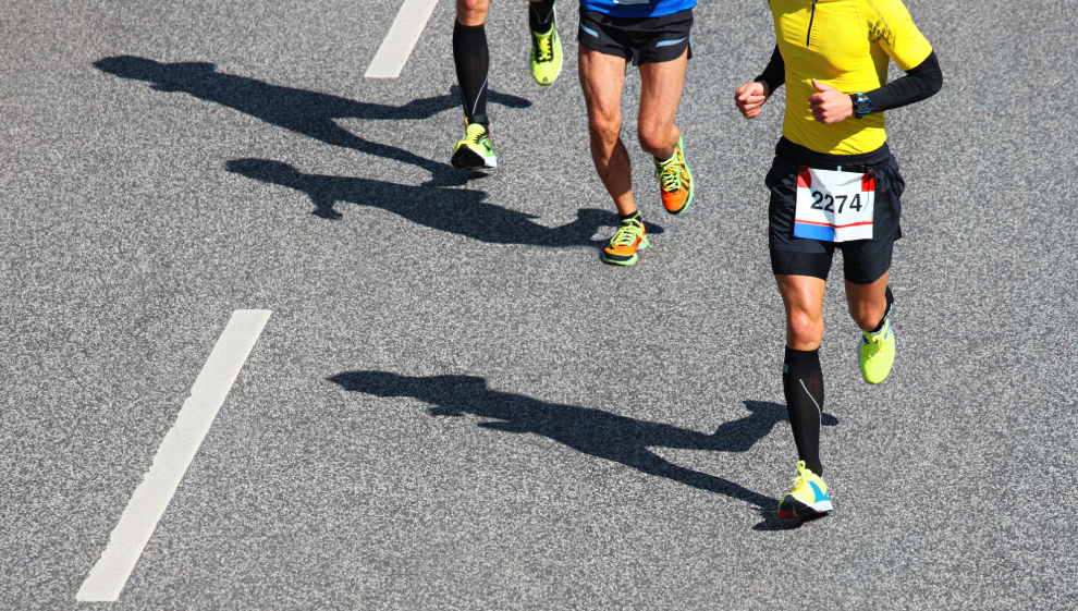 Marathon runners competing on a road race, wearing colorful athletic gear with race bibs, casting shadows on the asphalt.