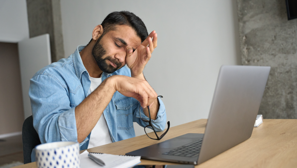 Tired man rubbing his eyes while working on a laptop, taking a break from remote work.