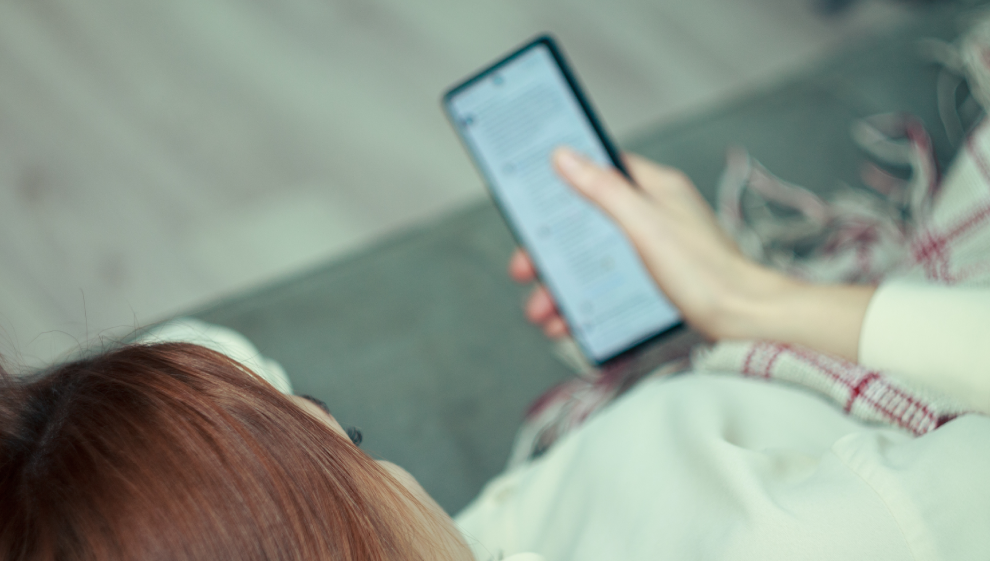 Woman holding and reading from a smartphone screen while wrapped in a cozy blanket on a couch.