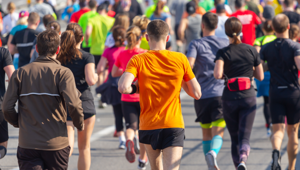 Large group of marathon runners seen from behind, participating in a city road race, with one runner in an orange shirt leading the pack.