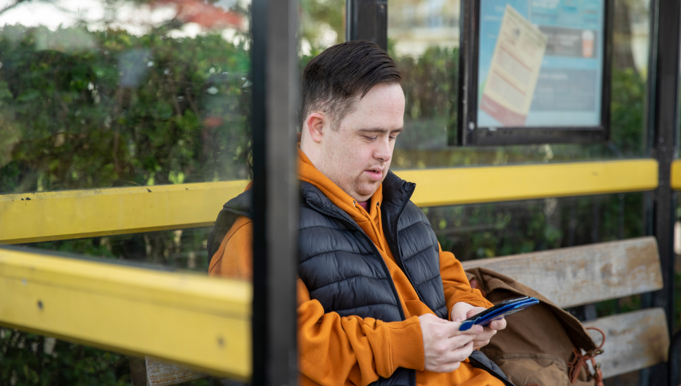 Man with Down syndrome sitting at a bus stop, wearing a black vest and orange hoodie, focused on his phone while waiting for public transportation.