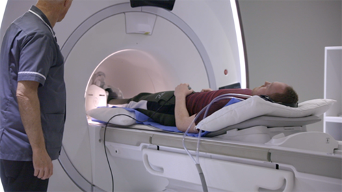 MRI Scan - Patient, lying on thie back, undergoing an MRI scan. A radiographer stands nearby.